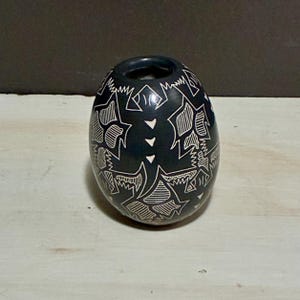 May include: A dark blue, egg-shaped vase with a wide opening. The vase is decorated with white, geometric patterns and shapes. The design includes star-like figures and linear elements. The vase is likely made of ceramic or stone.