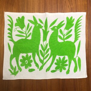 Puede incluir: Textil rectangular blanco con un diseño bordado en verde vibrante. El diseño presenta dos ciervos enfrentados, rodeados de flores y follaje estilizados. El bordado es detallado y el color es brillante.