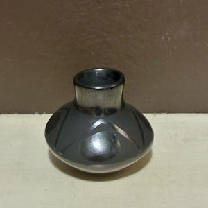 May include: A small, dark gray ceramic vase with a rounded body and a narrow neck. The vase has a glossy finish and is decorated with geometric patterns. The vase is sitting on a light-colored surface.