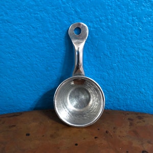 Op de afbeelding: Een zilveren metalen lepel met een gehamerde afwerking en een afgeronde steel. De steel heeft een klein gaatje bovenaan.