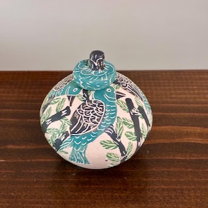 Op de afbeelding: Een ronde keramische pot met deksel op een matte crèmekleurige basis, voorzien van een grafisch patroon van zwarte en turquoise vogels op takken. Het deksel heeft een kleine, ronde knop en volgt het vogelmotief van de pot zelf.