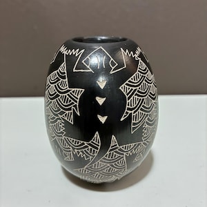 May include: A small, egg-shaped black vase featuring intricate white etched designs of a lizard or gecko. The surface is smooth and matte, with the creature's body centred and surrounded by geometric, wave-like patterns carved into the dark clay.