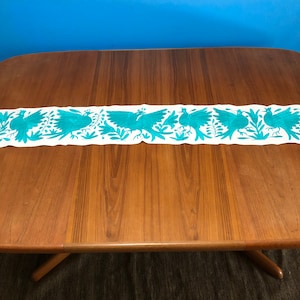 May include: A white table runner with a teal blue embroidered design of birds and flowers. The runner is on a wooden table.