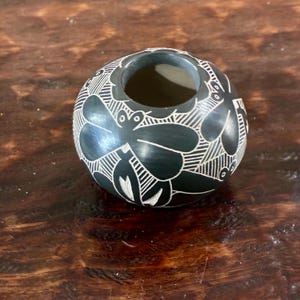 May include: A dark gray, spherical ceramic vase with a wide opening. The vase is decorated with a white, etched floral pattern. The vase is sitting on a wooden surface.