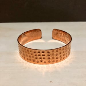 Op de afbeelding: Een koperen armband met een gehamerde textuur. De armband is een eenvoudige, massieve band met een gladde, afgeronde rand.
