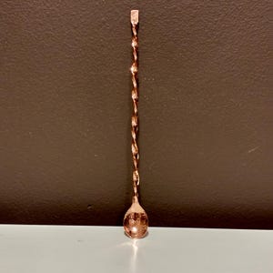 May include: A copper-colored bar spoon with a twisted stem and a teardrop-shaped bowl. The spoon has a flat, rectangular end for muddling. The design is sleek and functional, suitable for mixing cocktails.
