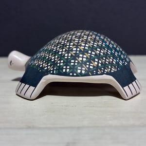 May include: A ceramic turtle figurine with a patterned shell. The shell has a dark blue background with a repeating pattern of green, white, and yellow geometric shapes. The turtle's head and feet are light pink, and the underside is white.