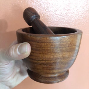 May include: A wooden mortar and pestle set. The mortar is a bowl shape with a rounded base, and the pestle is a cylindrical shape with a rounded top. Both pieces are made of a dark brown wood with visible grain.