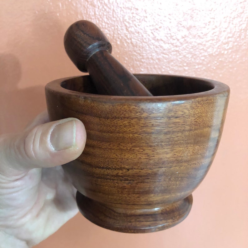 May include: A wooden mortar and pestle set. The mortar is a bowl shape with a rounded base, and the pestle is a cylindrical shape with a rounded top. Both pieces are made of a dark brown wood with visible grain.