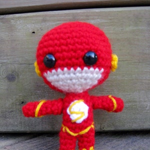 Puede incluir: Una muñeca de superhéroe de crochet roja y amarilla con un rayo blanco en el pecho. La muñeca tiene ojos negros y una boca blanca.