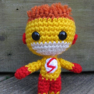 Puede incluir: Una figura de juguete de crochet del Flash, un superhéroe con supervelocidad. La figura es amarilla con detalles rojos y un rayo blanco en el pecho. La figura tiene el pelo rojo y naranja y ojos negros.
