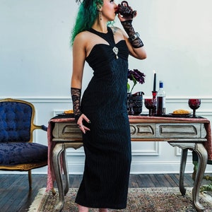 Puede incluir: Una mujer con el pelo verde y un vestido negro está de pie frente a una mesa con una copa de vino tinto. La mesa está puesta con un mantel rojo, velas y otras copas. La mujer lleva un sombrero negro y guantes.