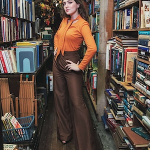 Puede incluir: Una mujer con una camisa de manga larga de color naranja brillante con cuello alto y un pantalón marrón de piernas anchas se encuentra en una librería. Las estanterías están llenas de libros.