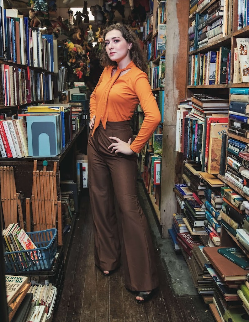May include: A woman wearing a bright orange long-sleeved shirt with a high neck and a brown pair of wide-leg pants stands in a bookstore. The shelves are filled with books.