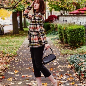 Op de afbeelding: Een vrouw in een geruit jasje, zwarte broek en beige hakken loopt over een pad bedekt met herfstbladeren. Ze draagt een zwarte handtas.