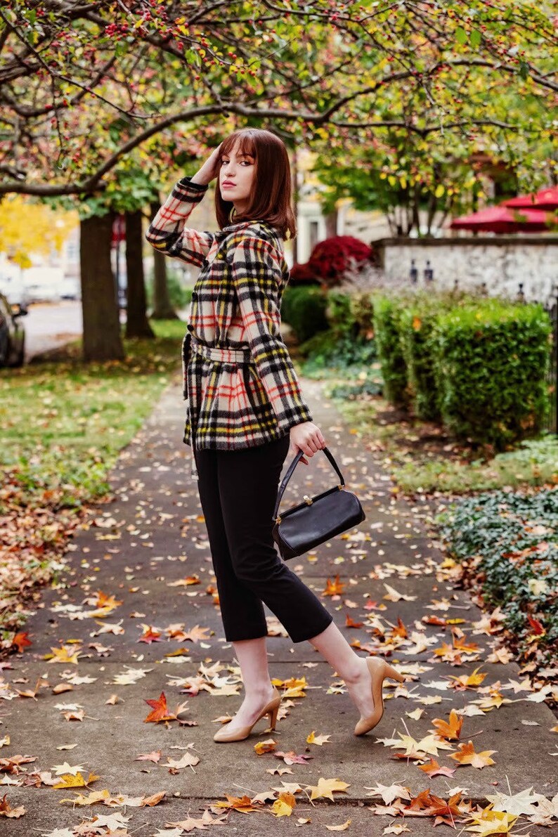 Op de afbeelding: Een vrouw in een geruit jasje, zwarte broek en beige hakken loopt over een pad bedekt met herfstbladeren. Ze draagt een zwarte handtas.