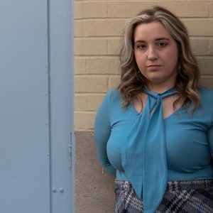 May include: A woman with long, wavy, light brown hair is wearing a light blue blouse with a large bow tie. She is standing in front of a brick wall and a blue door.