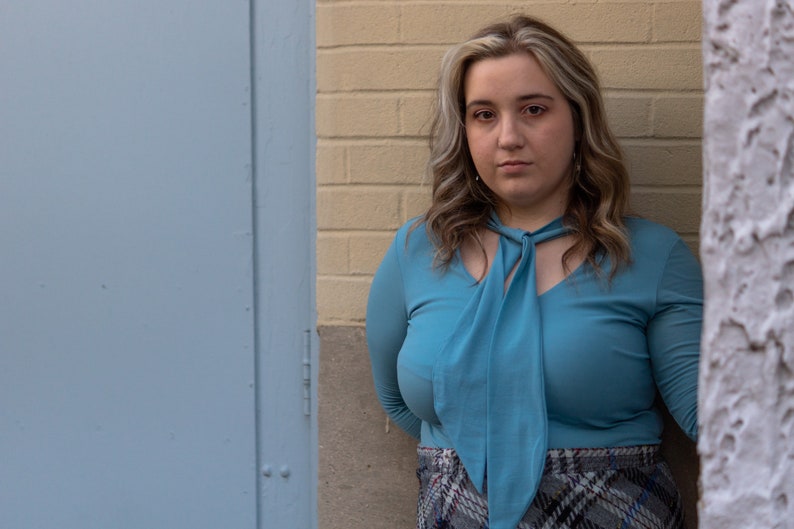 May include: A woman with long, wavy, light brown hair is wearing a light blue blouse with a large bow tie. She is standing in front of a brick wall and a blue door.