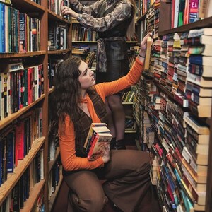 May include: Two people browse bookshelves in a bookstore. One person is wearing a brown and orange outfit and holding a stack of books. The other person is wearing a black and white patterned dress and is reaching for a book on a high shelf.