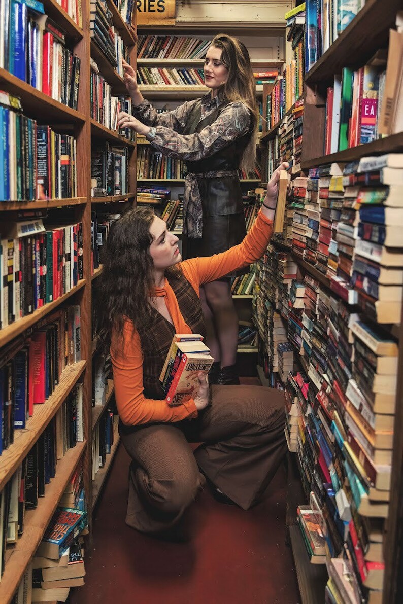 May include: Two people browse bookshelves in a bookstore. One person is wearing a brown and orange outfit and holding a stack of books. The other person is wearing a black and white patterned dress and is reaching for a book on a high shelf.