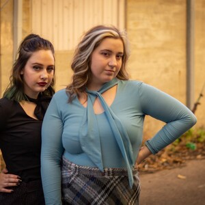 May include: Two women standing in front of a wooden wall. The woman on the left is wearing a black long-sleeved shirt with a black bow tie. The woman on the right is wearing a light blue long-sleeved shirt with a light blue bow tie and a plaid skirt.
