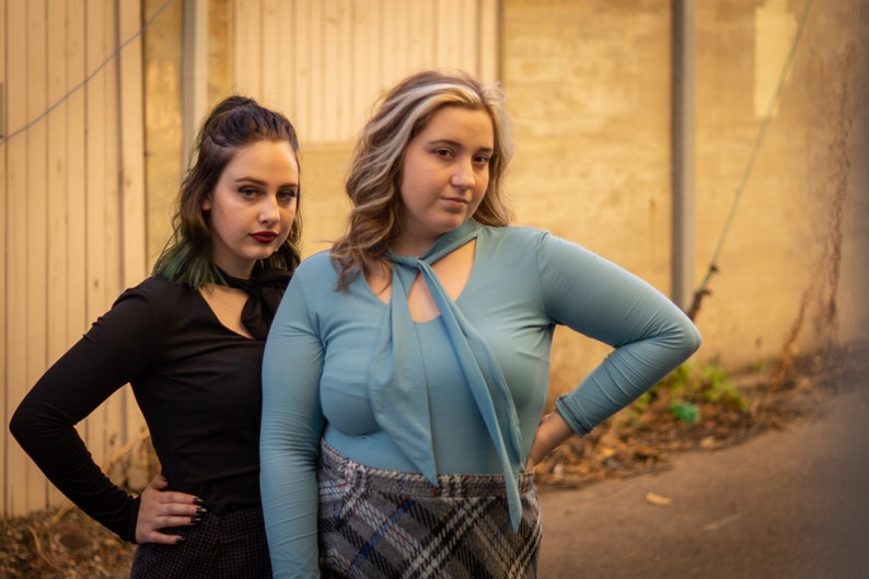 May include: Two women standing in front of a wooden wall. The woman on the left is wearing a black long-sleeved shirt with a black bow tie. The woman on the right is wearing a light blue long-sleeved shirt with a light blue bow tie and a plaid skirt.