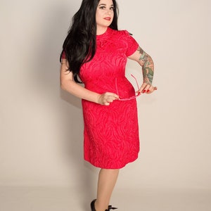 May include: A woman wearing a red dress with a textured pattern and black heels. She is holding a pair of pink glasses.