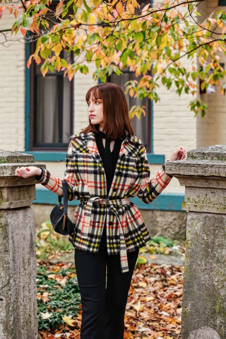 Op de afbeelding: Een vrouw draagt een zwarte turtleneck en een geruit jasje met een zwarte riem. Ze staat voor een stenen pilaar en een huis met herfstbladeren op de achtergrond.