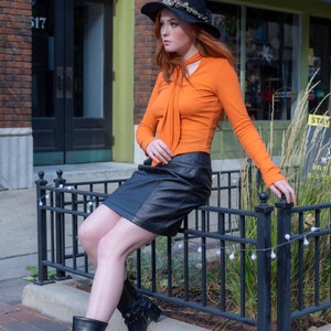May include: A woman wearing a black witch hat with a jeweled embellishment, an orange long-sleeved shirt, a black leather skirt, and black boots with silver accents. She is sitting on a black metal fence with white lights.