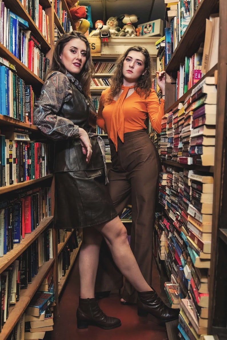 May include: Two women standing in a bookstore, one wearing a brown leather skirt and a patterned blouse, the other wearing a bright orange top and brown pants.
