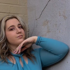 May include: A woman with long blonde hair is wearing a light blue, sheer, long-sleeved blouse with a ruffled neckline. She is leaning against a wall with her hand on her chin.