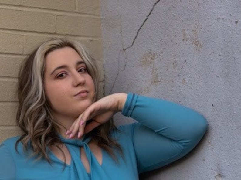 May include: A woman with long blonde hair is wearing a light blue, sheer, long-sleeved blouse with a ruffled neckline. She is leaning against a wall with her hand on her chin.