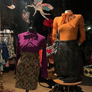 May include: Two mannequins wearing different outfits. The mannequin on the left is wearing a purple top with a bow and a colorful patterned skirt. The mannequin on the right is wearing an orange top with a bow and a black skirt.