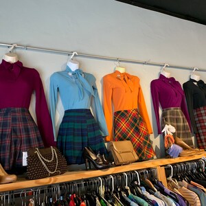 May include: Five different outfits on hangers, each with a long-sleeved blouse and a plaid skirt. The blouses are in shades of purple, blue, orange, and black. The skirts are in various shades of red, green, and black.