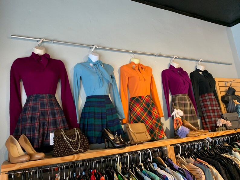 May include: Five different outfits on hangers, each with a long-sleeved blouse and a plaid skirt. The blouses are in shades of purple, blue, orange, and black. The skirts are in various shades of red, green, and black.