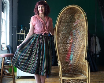 May include: A woman wearing a pink blouse and a black and multi-colored striped skirt stands next to a wicker peacock chair. The chair has a high back and a woven seat.