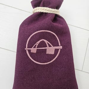 May include: A deep purple drawstring bag with a light pink embroidered design. The bag is made of a textured fabric and is tied at the top with a thick, cream-colored rope. The embroidered design is a stylized logo.