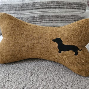Op de afbeelding: Een botvormig honden kussen in een bruinachtige stof met een zwart silhouet van een teckel. Het kussen ligt op een grijze, getextureerde ondergrond, met een gestreept kussen op de achtergrond.