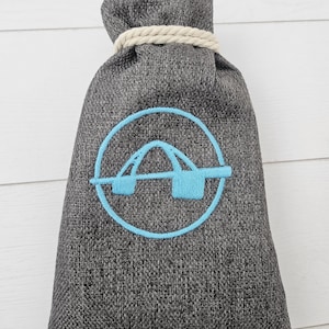 May include: A gray fabric bag with a light blue embroidered logo of a bridge inside a circle. The top of the bag is cinched with a beige rope. The bag is made of a textured material.