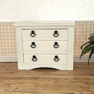 May include: A small white wooden chest of drawers featuring three horizontal drawers. Each drawer is equipped with two black metal drop-style pulls. The piece has a rectangular top surface and a slightly arched base design at the bottom front.