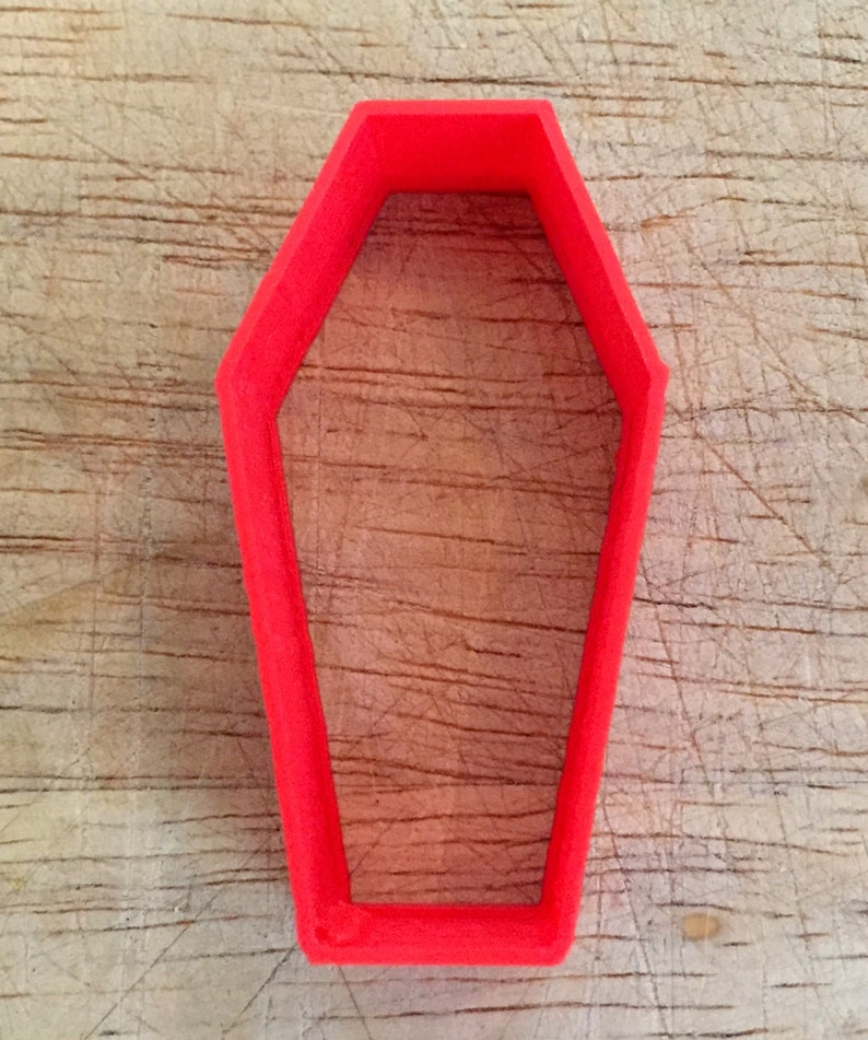 May include: Bright red coffin-shaped cookie cutter. The cutter is made of plastic and has a simple, geometric design. Perfect for Halloween-themed baking or crafting projects. The cutter is shown on a wooden surface.