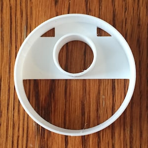 May include: A white plastic cookie cutter with a circular outer shape and a smaller circular cutout in the center. Two triangular cutouts are on the top edge. The cutter sits on a wooden surface.