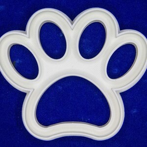 May include: A white paw print-shaped object, likely a cookie cutter or mold, against a blue background. The paw print has five rounded lobes, with a raised, rounded border. The object is made of a solid, opaque material.