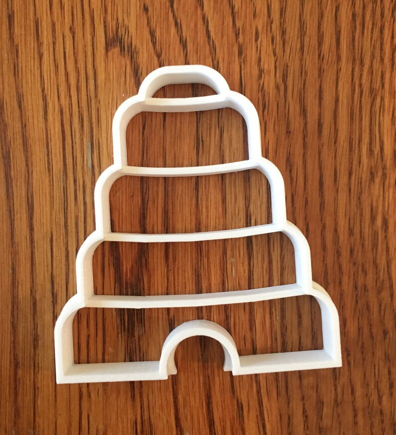May include: A white plastic cookie cutter in the shape of a tiered beehive. The cutter has a rounded top, three horizontal tiers, and a curved opening at the bottom. The cutter is on a wooden surface.
