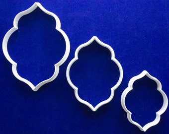 May include: Three white plastic cookie cutters in an arabesque shape, arranged from large to small. The cutters are on a solid blue background. These cutters are ideal for creating cookies or other treats in a unique shape.