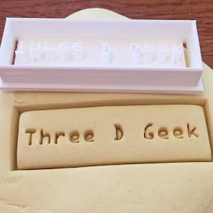 May include: A white plastic stamp with the letters "LUL66 D R66K" is positioned above a rectangular impression in a light yellow clay. Below, the words "Three D Geek" are imprinted in the clay. The image showcases a crafting tool for creating custom text.