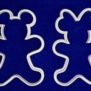 May include: Two white metal cookie cutters in the shape of teddy bears. One has rounded ears, the other has a bow-shaped head. The cutters are on a dark blue background. Ideal for baking themed cookies.
