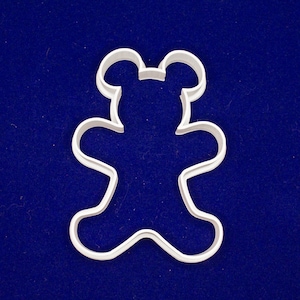 May include: A white plastic teddy bear cookie cutter. The cutter is shaped like a classic teddy bear with rounded features, including a head with ears, arms, and legs. The cutter is set against a dark blue background.