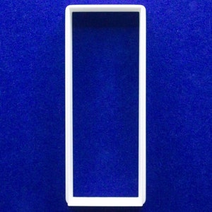 May include: A white rectangular cookie cutter with a hollow center, set against a deep blue background. The cutter's shape is a simple rectangle, perfect for creating uniform shapes in baking.