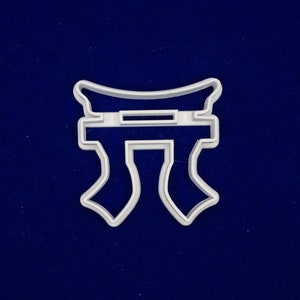 May include: A white plastic cookie cutter shaped like a Japanese Torii gate, on a dark blue background. The cutter features a rectangular top with a horizontal opening and two curved pillars.